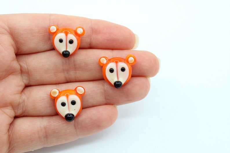 May include: Three orange glass animal buttons with white faces and black eyes. The buttons are shaped like foxes.