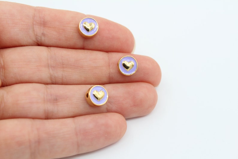 May include: Three gold-colored round beads with a lavender enamel coating and a gold heart design.