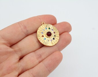 May include: Gold-toned pendant with a circular design featuring a red gemstone in the center surrounded by smaller colorful gemstones. The pendant has a raised edge and is inscribed with text.