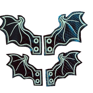 Op de afbeelding: Vier zwarte leren vleermuisvleugelpatches met een groene omtrek en stiksels. Elk patch heeft twee metalen lussen aan de achterkant.