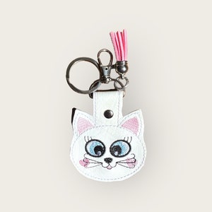 May include: White cat-shaped keychain with embroidered details. The cat has blue eyes, pink ears, and whiskers. A pink tassel hangs from the top. The keychain has a silver clasp and a snap closure.