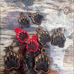 May include: Embroidered bear paw patches in brown, black, and red. The patches feature a detailed paw print with claws, and a loop at the top for sewing. The patches are arranged on a wooden surface.