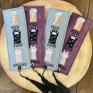 May include: Four rectangular bookmarks with black tassels. Each bookmark features a Rock, Paper, Scissors design with a paw print and text. The bookmarks are in shades of gray and burgundy, with a light brown wooden background.