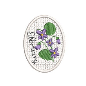 Könnte beinhalten: Ein weißes ovales Stickmuster mit einem violetten und grünen Blumendesign und dem Wort "Februar" vertikal in Kursivschrift geschrieben.