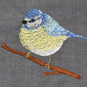 Op de afbeelding: Een geborduurde blauwe vogel op een bruine tak. De vogel heeft een blauwe kop en vleugels, een gele buik en witte accenten.