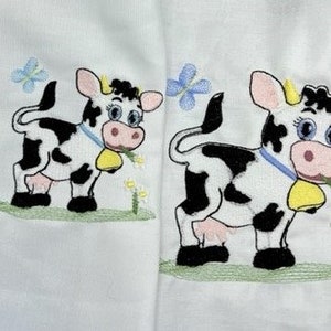 May include: Two embroidered white towels with black and white cow designs. Each cow is holding a flower in its mouth and has a bell around its neck. The towels have a blue flower design in the top corner.