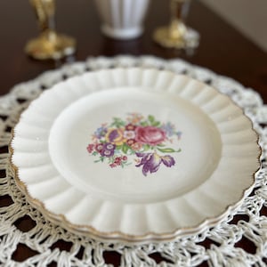 May include: A white ceramic dinner plate with a gold rim and a floral design in shades of pink, purple, yellow, and green. The plate is on a white crocheted doily.