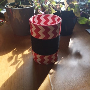 Può includere: Un nastro rosso e bianco a chevron avvolto attorno a un cilindro nero. Il cilindro è appoggiato su una superficie di legno.