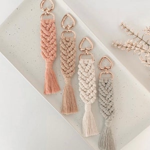 May include: Four handmade macrame keychains with heart-shaped gold metal clasps and tassels. The keychains are in shades of pink, beige, white, and grey.