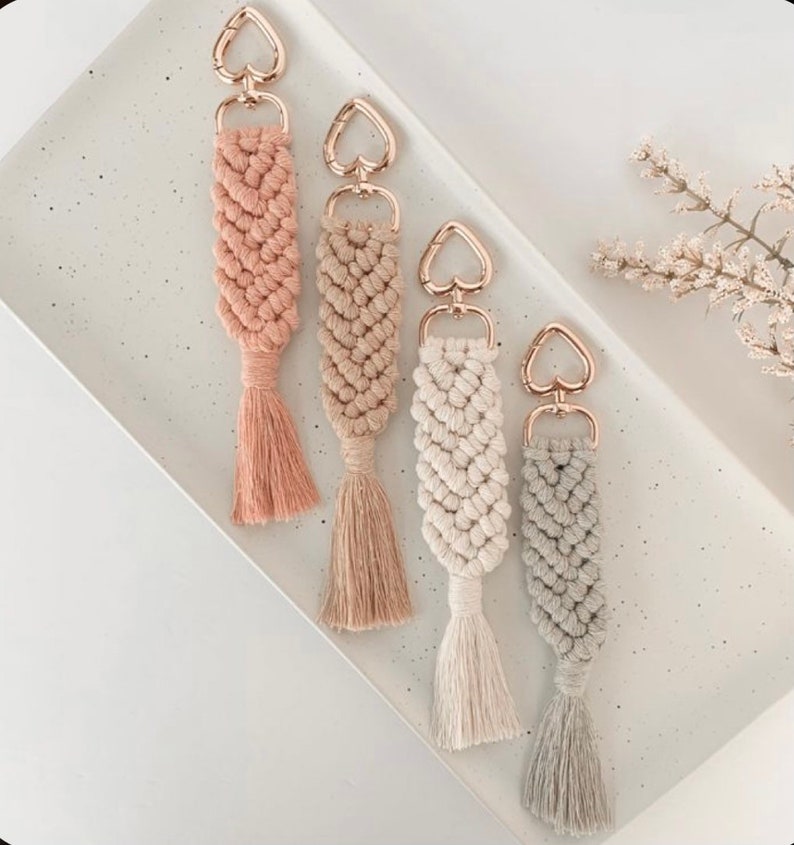 May include: Four handmade macrame keychains with heart-shaped gold metal clasps and tassels. The keychains are in shades of pink, beige, white, and grey.