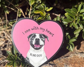 May include: A pink heart-shaped sticker with a black border. The sticker features a smiling dog illustration with a bandana that says "BLIND DOG." Above the dog is the text "I see with my heart."