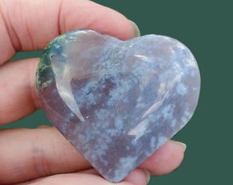 May include: A heart-shaped stone with a mix of colors, including blue, purple, and green. The stone has a polished surface and a natural, organic appearance. The heart is held in a hand against a green background.