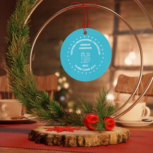 May include: A blue round ornament with white text that reads "Certified Professional Controller, Andrew Anderson, Flying Cloud ATCT, 2023". The ornament is hanging from a red ribbon.