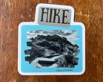 May include: A blue and white sticker with a black and white photo of a hiking trail with the text "HIKE." and "Chaos Caravan" at the bottom.