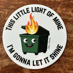 Peut inclure: Un autocollant rond blanc avec du texte noir qui dit "This little light of mine, I'm gonna let it shine." Une poubelle verte avec un visage souriant et une flamme jaune sur le dessus est au centre de l'autocollant.