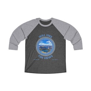 以下が含まれることがあります： 濃いグレーと薄いグレーのベースボールTシャツに、青い円形のグラフィックがプリントされています。グラフィックには、湖のある山岳風景と「WILL HIKE FOR VIEWS」というテキストが描かれています。