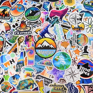 May include: A collection of colorful stickers with various designs, including mountains, a compass, a campfire, a peace sign, and the words "Adventure", "Good Vibes", and "Live Free".