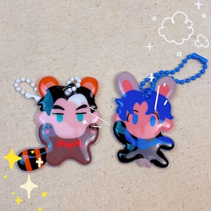 Könnte beinhalten: Zwei bunte Schlüsselanhänger mit Cartoon-Charakteren. Ein Schlüsselanhänger ist ein Waschbär mit einer roten Fliege und einem schwarz-orange gestreiften Schwanz. Der andere Schlüsselanhänger ist ein blauer Hase mit einem blauen Schal. Beide Schlüsselanhänger sind aus klarem Kunststoff mit glänzender Oberfläche.