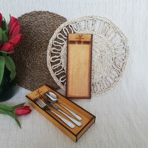 May include: A wooden cutlery set with a fork, spoon, and knife in a rectangular box. The box is made of light brown wood with a dark brown trim. A matching wooden holder is also present. Red tulips in a green vase and woven placemats complete the scene.