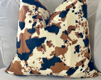 May include: A decorative throw pillow with a brown, black, and white cowhide print. The pillow is made of a soft, plush fabric.