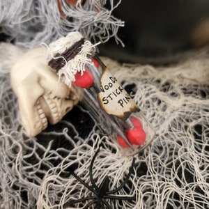 Peut inclure: Une petite bouteille en verre avec un bouchon en liège et une étiquette qui indique "Voodoo Stick Pins". La bouteille est remplie d'épingles rouges et repose sur une toile d'araignée blanche.