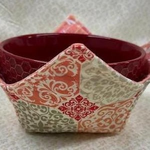 May include: A red bowl with a patterned fabric bowl cozy. The cozy is made of a patchwork of beige, pink, and red fabric with a floral design.