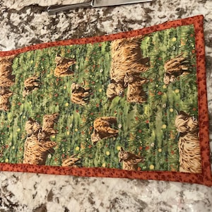 May include: Rectangular placemat featuring a repeating pattern of brown Highland cows on a green background with small red and yellow flowers. The placemat has a rust-colored border.