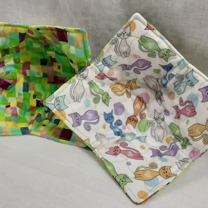 May include: Two quilted pot holders, one with a green and brown patchwork design and the other with a white background and a repeating pattern of colorful cats.