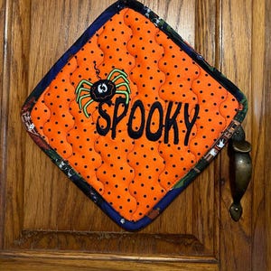 May include: Orange and black quilted pot holder with a spider and the word "SPOOKY" embroidered on it.