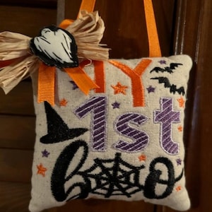 May include: A white fabric pillow with the words "My 1st Boo" embroidered in black and purple. The pillow is decorated with a black witch's hat, a spider web, and bats. It has a orange ribbon and a straw bow with a white ghost.