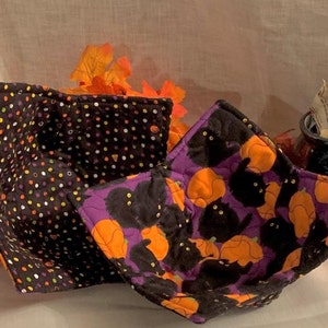 May include: Two quilted bowl cozies. One is black with white and orange polka dots. The other is purple with black cats and orange pumpkins.