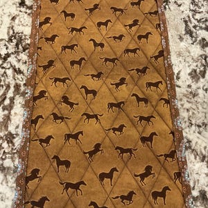 May include: A rectangular, quilted placemat featuring a repeating pattern of brown horse silhouettes on a tan background. The edges are trimmed with a patterned border in shades of brown, blue, and white. The placemat has a diamond-stitched pattern.