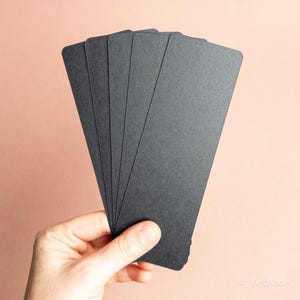 May include: A hand holding a fanned stack of six black rectangular cards with rounded corners. The cards have a textured surface and are arranged against a soft peach-colored background. The cards are likely for a game or a craft project.