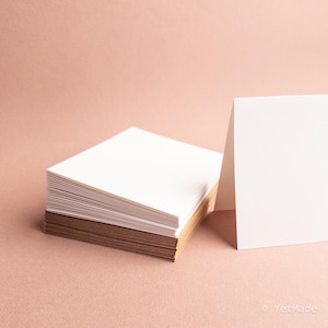 May include: A stack of white blank cards with a brown edge, and a single card standing upright. The cards are on a light pink surface. The cards are likely for invitations or notes.
