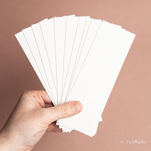 May include: A hand holding a fan of blank, white rectangular cards. The cards are fanned out, with the edges visible. The background is a solid, muted reddish-brown color. The cards are likely for notes or invitations.