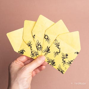 May include: A hand holding a fan of five yellow envelopes. Each envelope has a black bee pattern on the front. The background is a solid, muted, reddish-brown color.