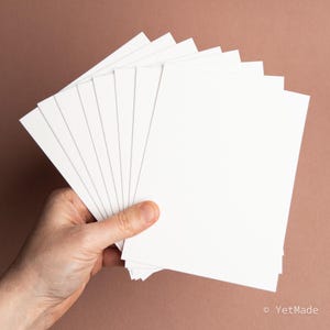 May include: A hand holds a fanned stack of blank, white rectangular cards against a brown background. The cards are thick and have a smooth surface, ideal for various creative projects.