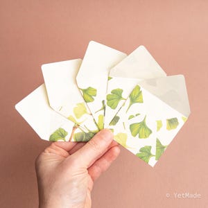May include: A hand holding several white envelopes with a ginkgo leaf pattern. The envelopes have a square shape and are open, revealing a blank interior. The ginkgo leaf design features green and yellow leaves on a white background. The background is a solid, muted brown.
