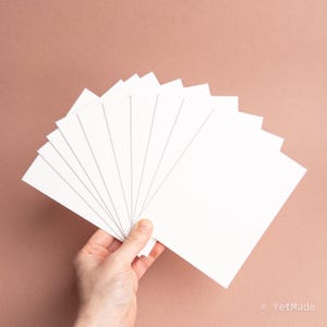 May include: A hand holding a fanned stack of blank, white rectangular cards against a solid, muted rose-colored background. The cards are evenly spaced, creating a fan-like arrangement. The image is well-lit, highlighting the smooth texture of the cards.