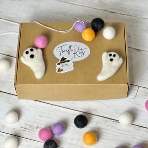 K&ouml;nnte beinhalten: Eine braune Pappschachtel mit Filz-Geisterdekorationen und bunten Filzkugeln. Auf der Schachtel befindet sich ein Aufkleber mit dem Text "Twilli Kits" und einem Cartoon-Geist. Die Filzkugeln sind in Rosa, Lila, Schwarz, Wei&szlig; und Orange gehalten.