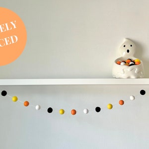 May include: A white shelf with a white ghost-shaped candy dish filled with orange, yellow, and black candy. A garland of orange, yellow, black, and white pom-poms hangs below the shelf. The text "LOOSELY SPACED" is written in white on an orange circle.