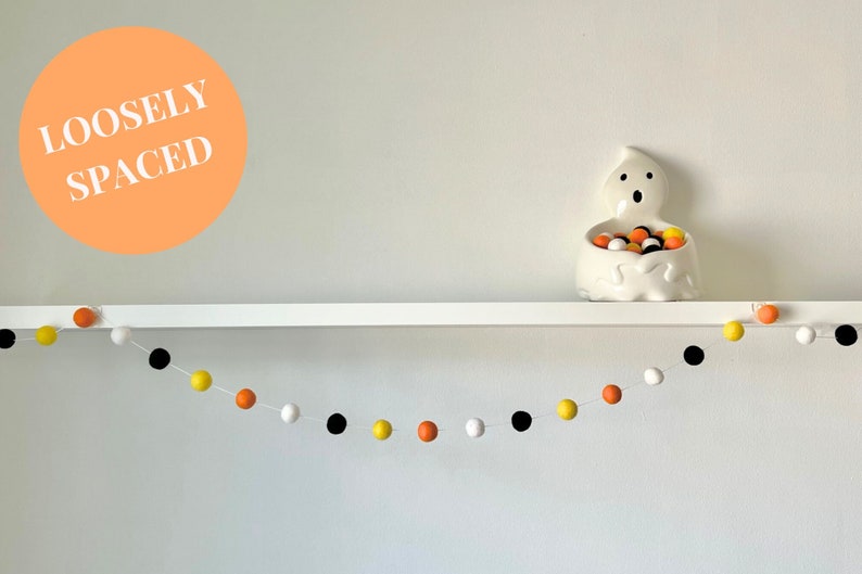 May include: A white shelf with a white ghost-shaped candy dish filled with orange, yellow, and black candy. A garland of orange, yellow, black, and white pom-poms hangs below the shelf. The text "LOOSELY SPACED" is written in white on an orange circle.