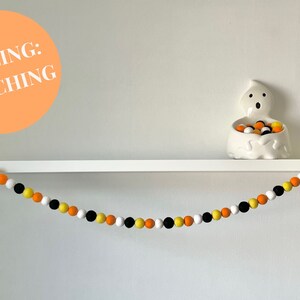 May include: A Halloween garland made of felt balls in orange, yellow, black, and white. The balls are touching each other and are strung on a white string. The garland is hanging on a white shelf with a white ghost figurine holding more felt balls.