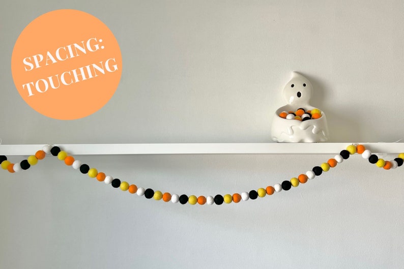 May include: A Halloween garland made of felt balls in orange, yellow, black, and white. The balls are touching each other and are strung on a white string. The garland is hanging on a white shelf with a white ghost figurine holding more felt balls.