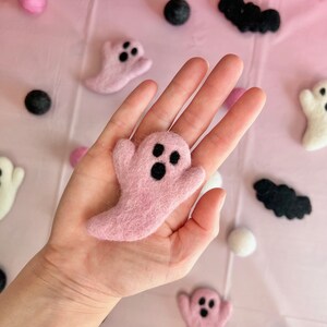 K&ouml;nnte beinhalten: Ein rosa Filzgespenst mit schwarzen Augen wird in einer Hand gehalten, umgeben von anderen Filzgespenstern, schwarzen Filzflederm&auml;usen und wei&szlig;en und rosa Filzkugeln. Die Objekte sind auf einer rosa Oberfl&auml;che angeordnet und schaffen ein festliches Halloween-Thema.