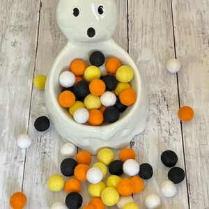 May include: A white ceramic ghost-shaped bowl filled with black, orange, and yellow felt balls. Some of the balls have spilled out of the bowl onto a white wooden surface.