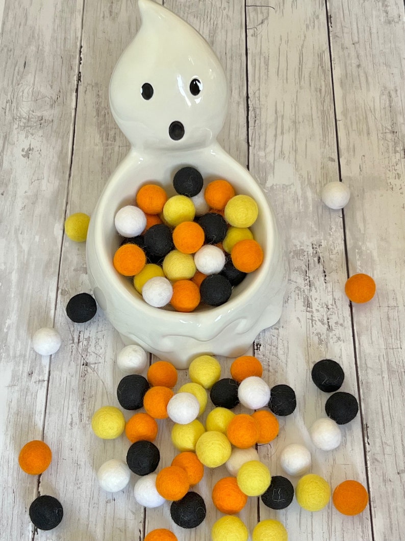 May include: A white ceramic ghost-shaped bowl filled with black, orange, and yellow felt balls. Some of the balls have spilled out of the bowl onto a white wooden surface.