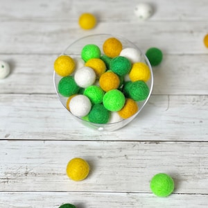 May include: A clear glass bowl filled with green, white, and yellow felt balls. The bowl is sitting on a white wooden surface.