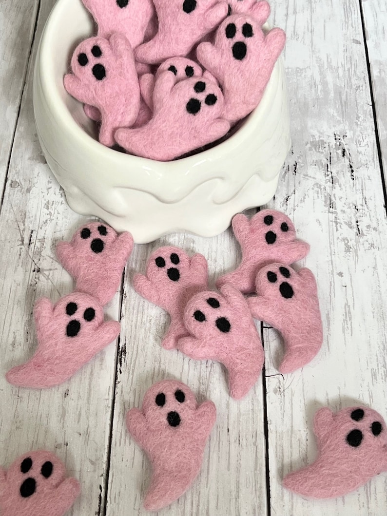 K&ouml;nnte beinhalten: Rosa Filz-Geisterdekorationen mit schwarzen Augen und M&uuml;ndern, einige in einer wei&szlig;en Schale. Diese handgefertigten Halloween-Ornamente sind auf einer hellen Holzoberfl&auml;che angeordnet. Ideal f&uuml;r Bastelarbeiten oder saisonale Dekoration.