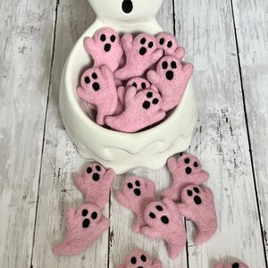 K&ouml;nnte beinhalten: Ein wei&szlig;er, geisterf&ouml;rmiger Keramikbeh&auml;lter gef&uuml;llt mit rosa Filz-Geisterfiguren. Jeder rosa Geist hat schwarze Augen und einen schwarzen Mund. Mehrere rosa Geister sind auf einer wei&szlig;en Holzoberfl&auml;che verstreut. Halloween-Dekoration.